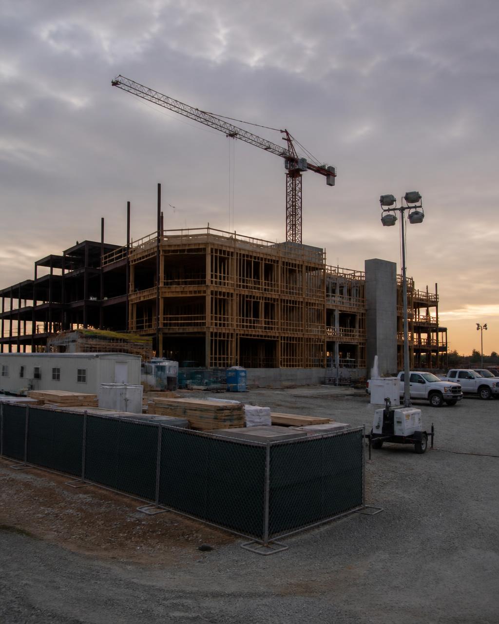 Fenced commercial construction site with jobsite trailers and a tower crane at dusk